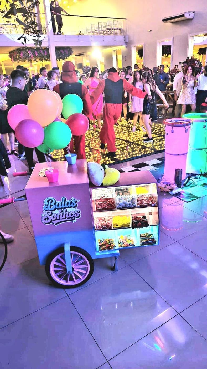 Carrinho Balas e Sonhos com balões coloridos ao lado da pista de dança iluminada, com convidados dançando ao fundo