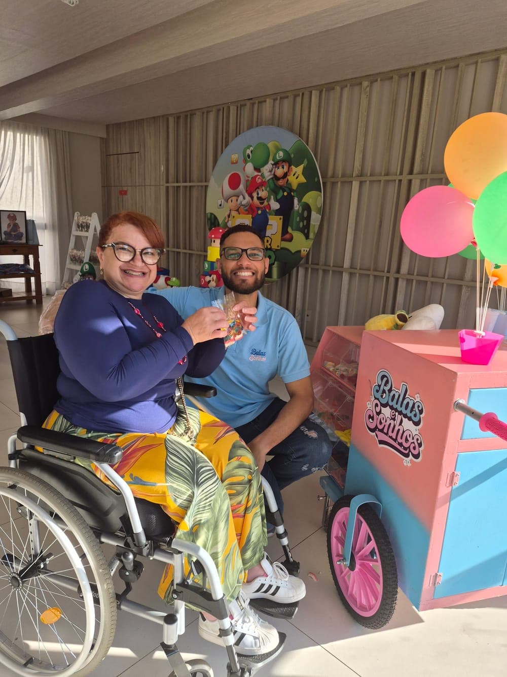 Convidada em cadeira de rodas segurando balas ao lado de um integrante da equipe, os dois sorrindo perto do carrinho