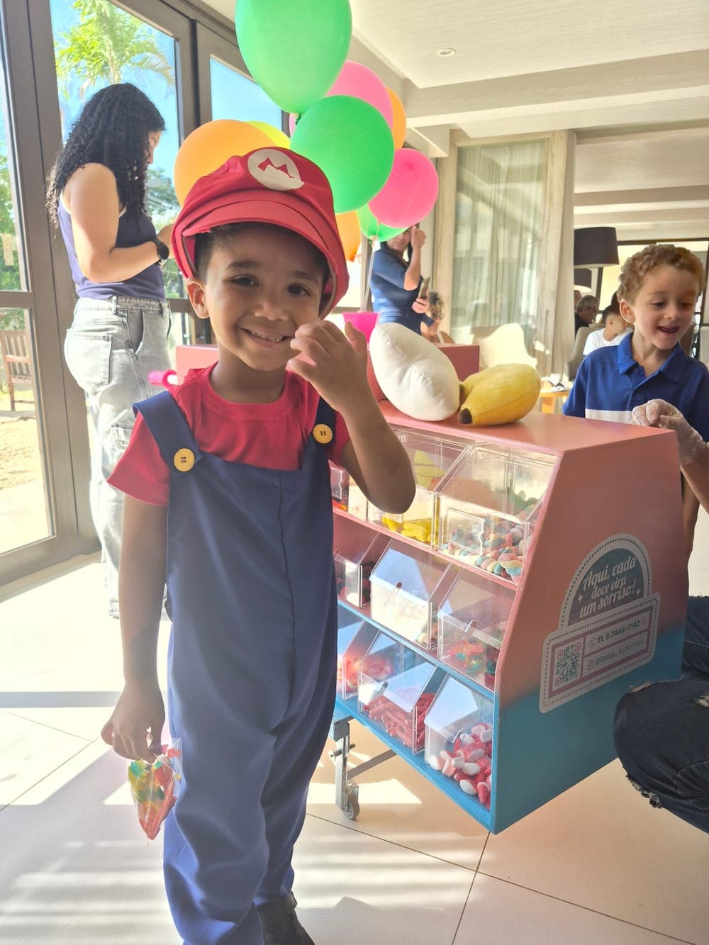 Menino fantasiado de Mario sorrindo com o saquinho de balas na mão, em frente ao carrinho Balas e Sonhos