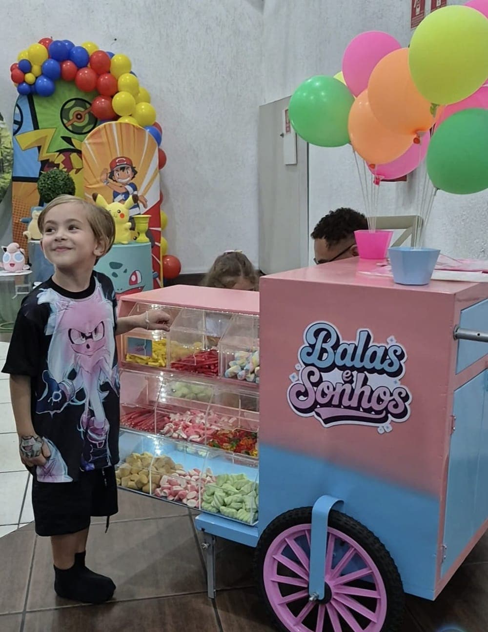 Menino sorrindo ao lado do carrinho Balas e Sonhos em uma festa com tema de Pokémon