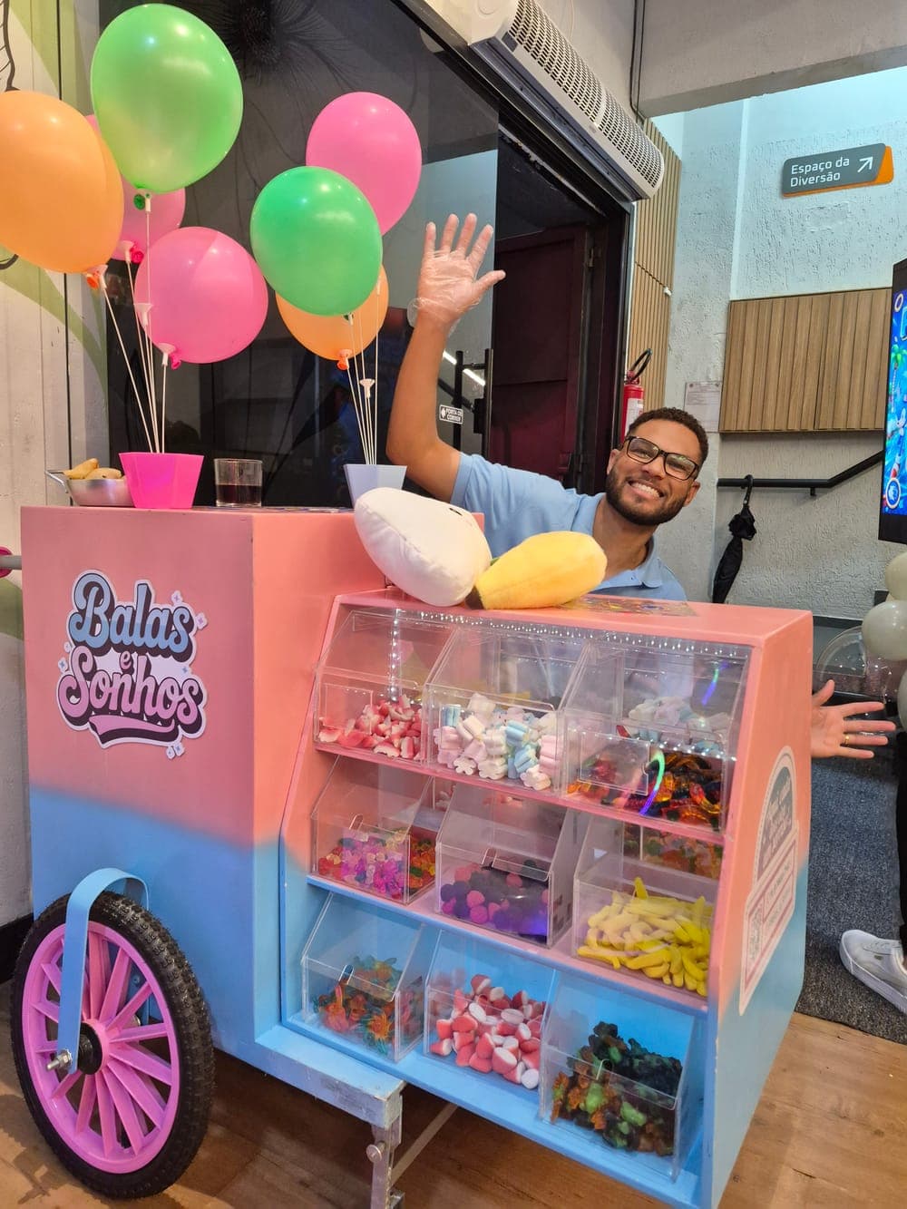 Integrante da equipe Balas e Sonhos sorrindo e acenando atrás do carrinho cheio de balas, com balões