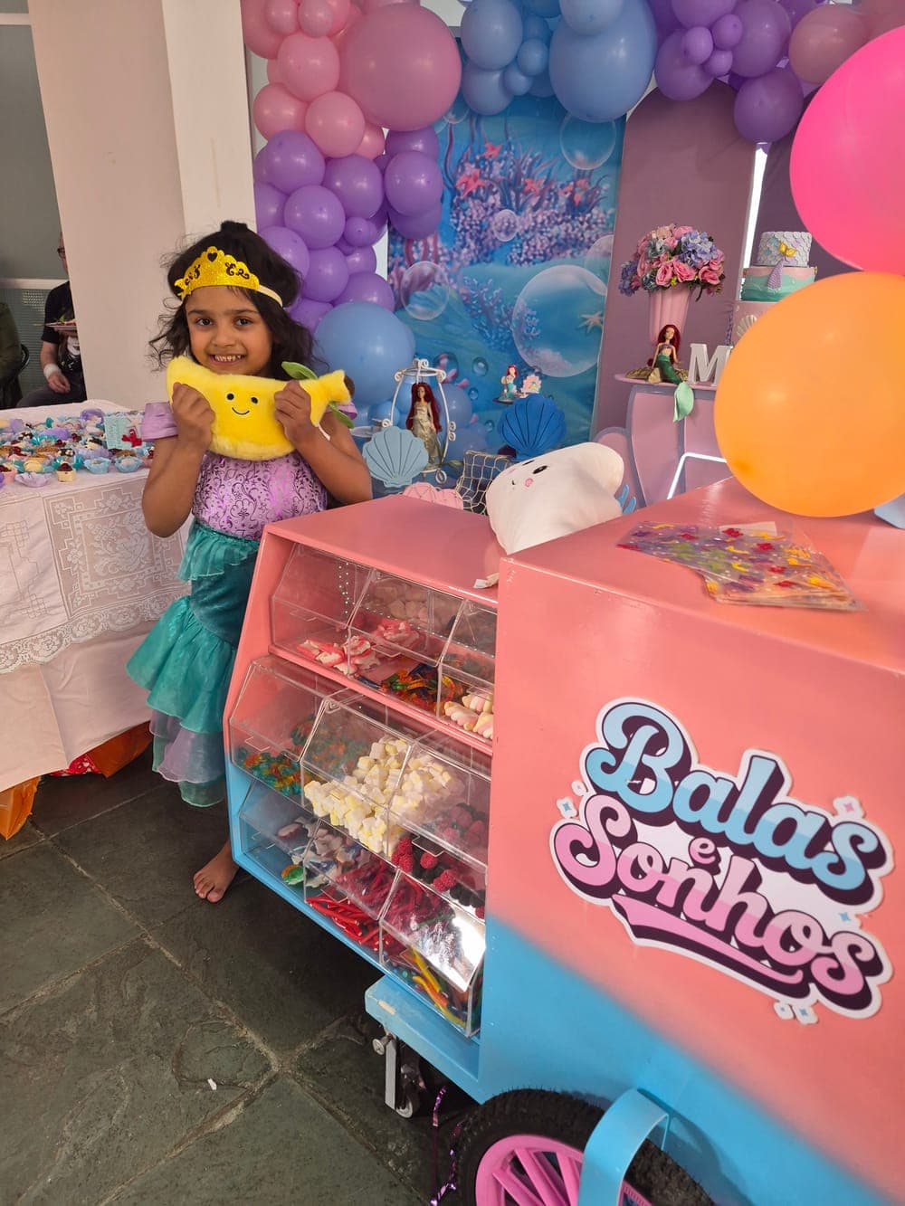 Menina fantasiada de sereia sorrindo e abraçando a pelúcia de banana ao lado do carrinho, em festa com tema de Pequena Sereia