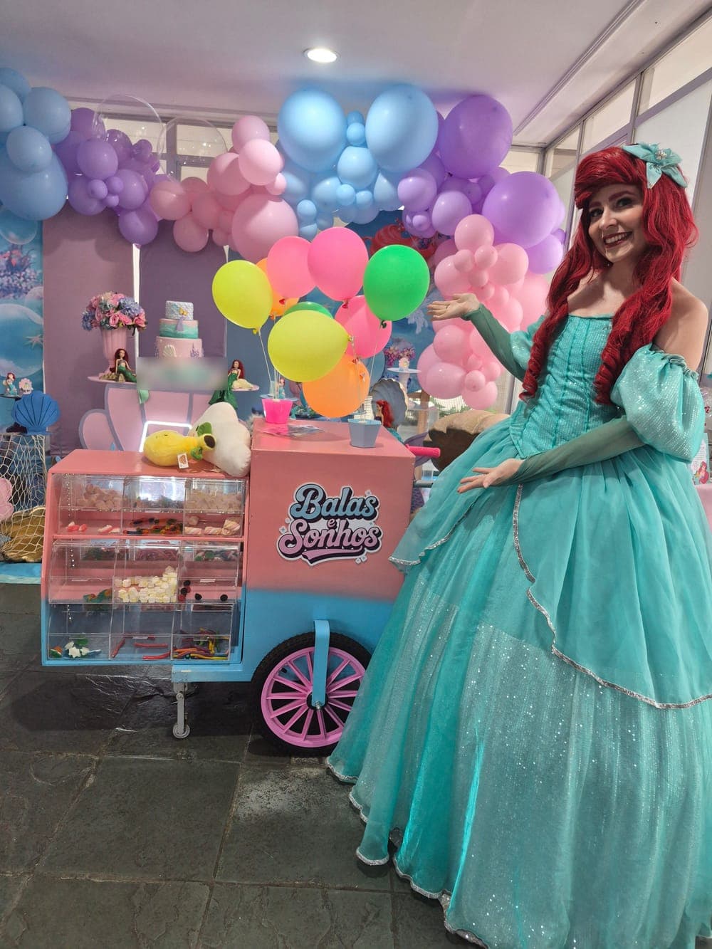 Carrinho Balas e Sonhos ao lado de uma personagem de Pequena Sereia, em festa decorada com balões em tons pastel
