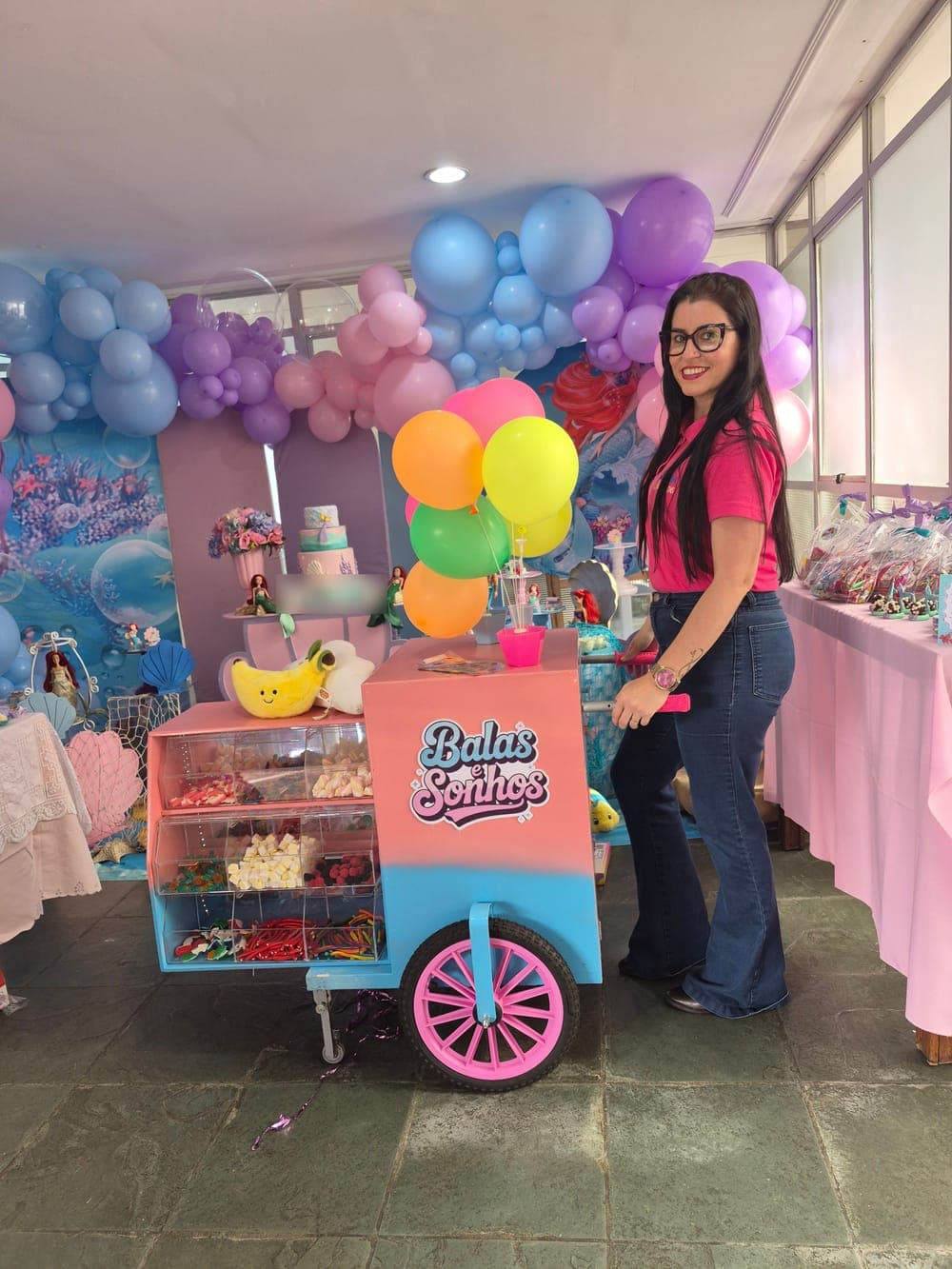 Priscilla, fundadora da Balas e Sonhos, de camisa rosa da marca, ao lado do carrinho em uma festa com tema de Pequena Sereia