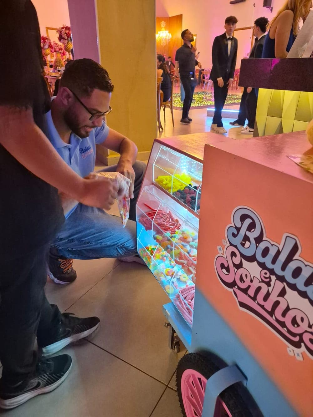 Integrante da equipe, de luvas, montando um saquinho de balas ao lado do carrinho iluminado, em uma festa de 15 anos