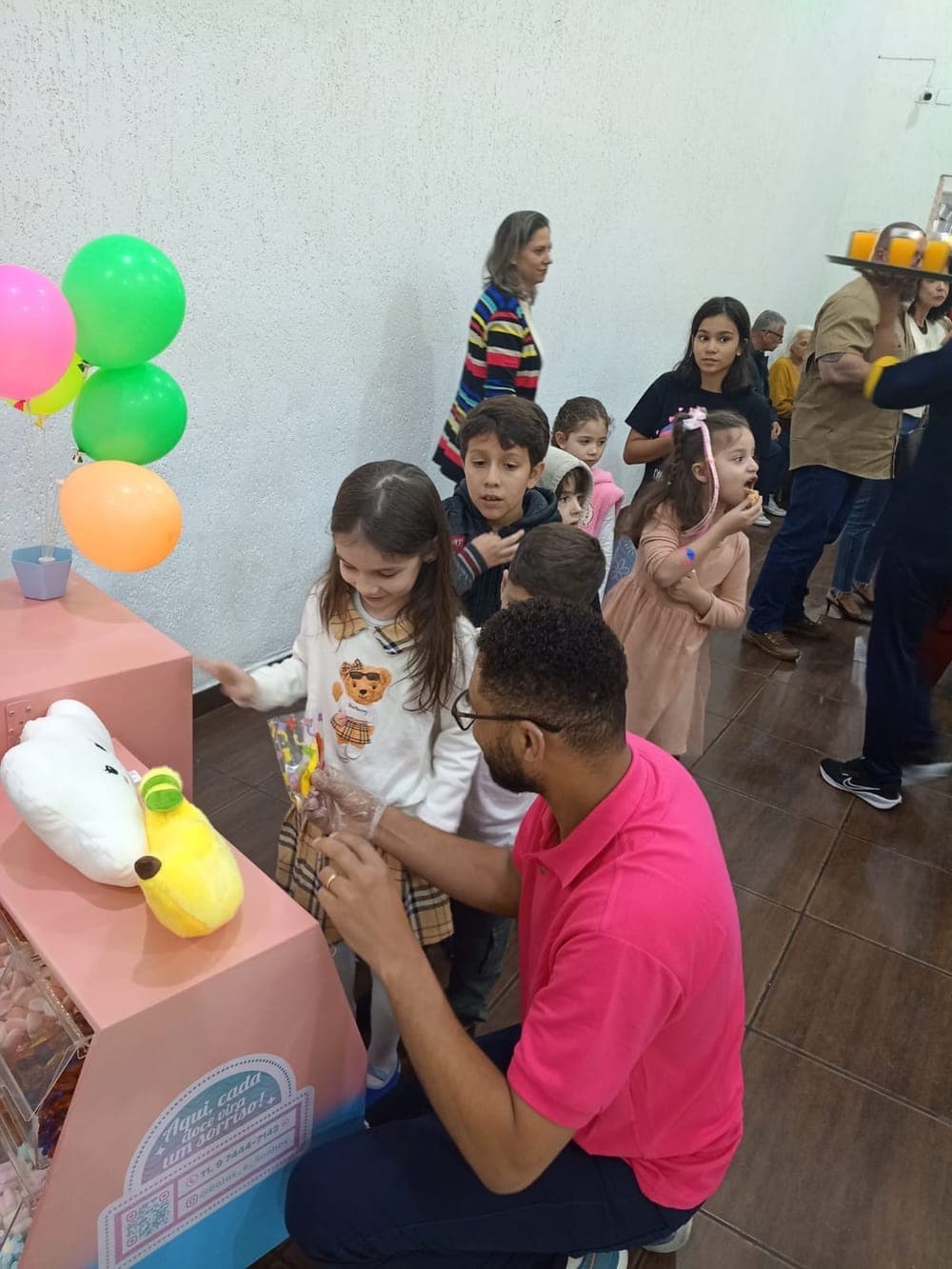 Integrante da equipe, de luvas, entregando o saquinho de doces para uma menina, com outras crianças na fila do carrinho